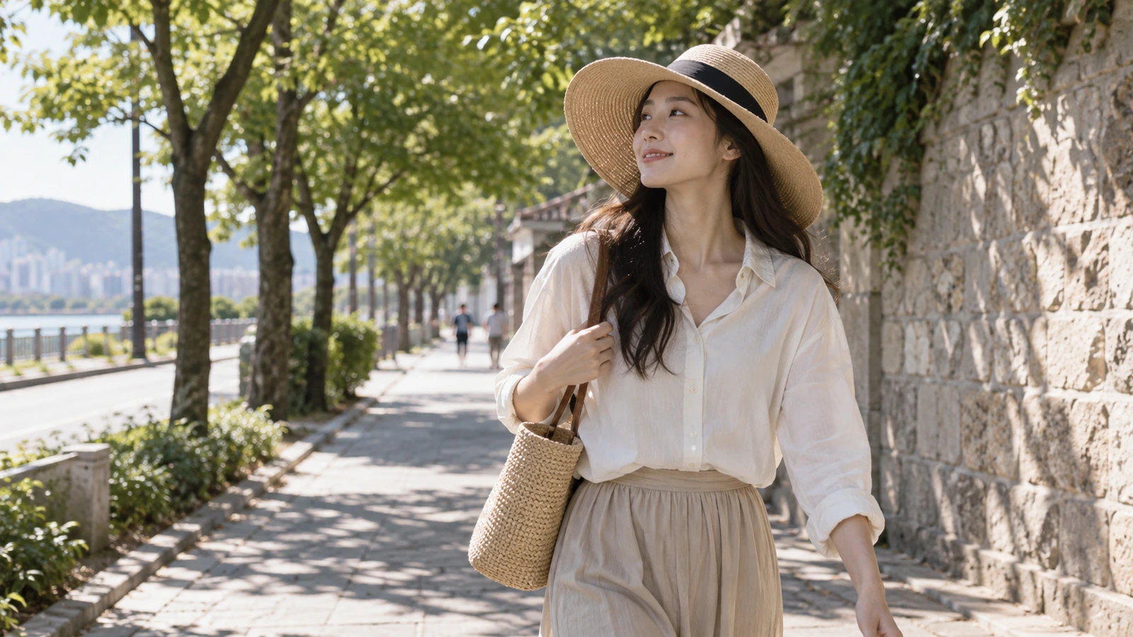 넓은 챙 모자를 쓰고 가방 끈을 잡은 채 나무 그늘이 드리운 길을 걷는 여성