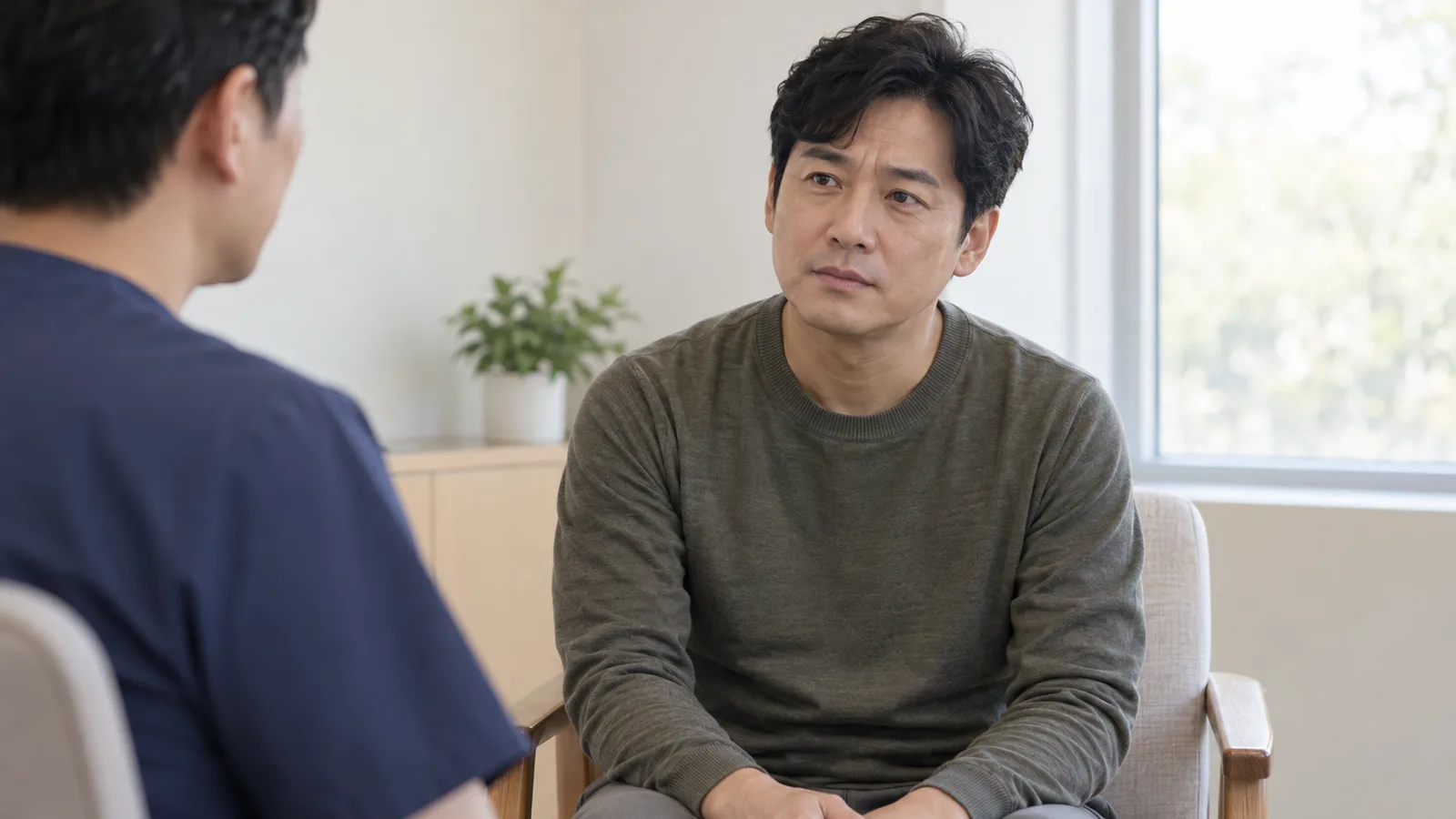 밝은 방에서 남색 의료복을 입은 사람의 뒷모습 너머로 의자에 앉은 중년 남성이 살짝 걱정스러운 표정으로 상대를 바라보고 있다.